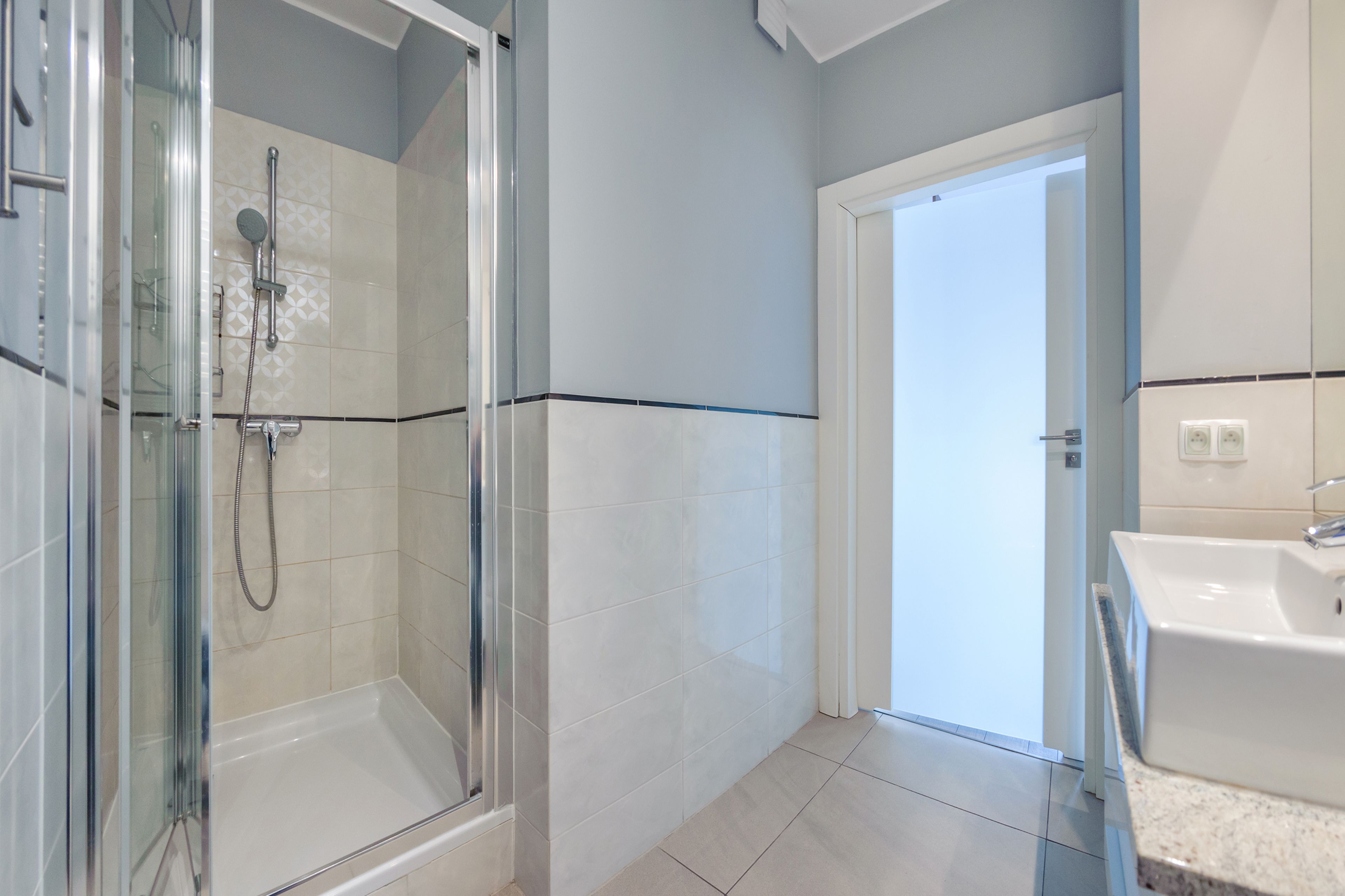 A modern bathroom with a glass-enclosed shower, a white sink, and a door leading to another room.