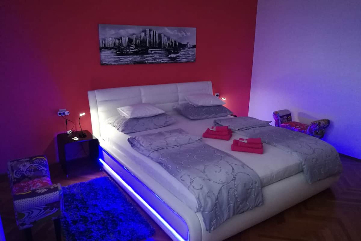 A modern bedroom with a large bed, a cityscape painting on the wall, and a blue ambient light strip along the bed's edge.