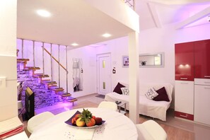 A modern living room with a staircase, a dining area with a fruit bowl, and a kitchen with red cabinets. - Apartments Ella (Split)