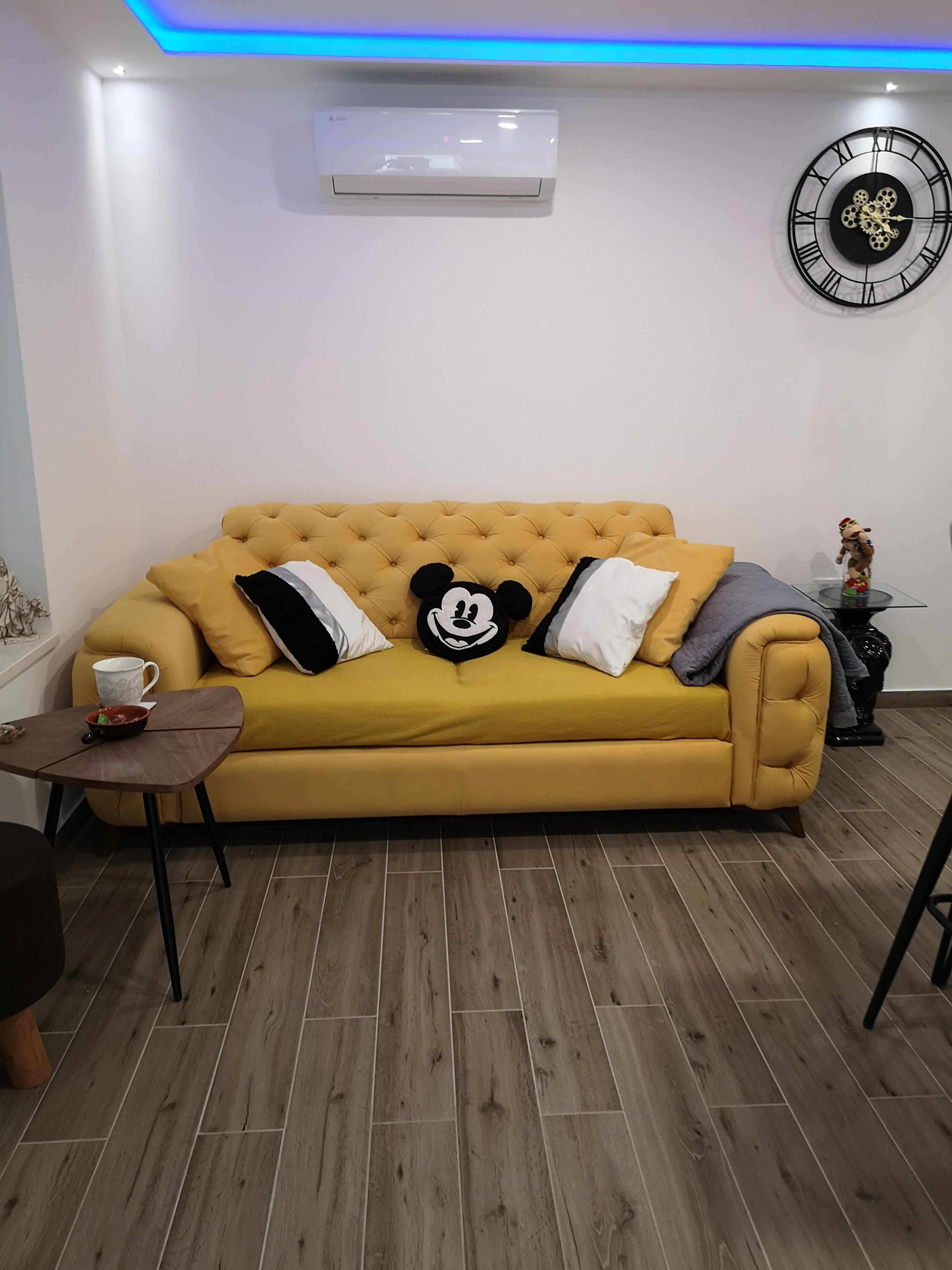 Een woonkamer met een geel sofa, een Mickey Mouse kussen, een koffiekop en een wandklok.