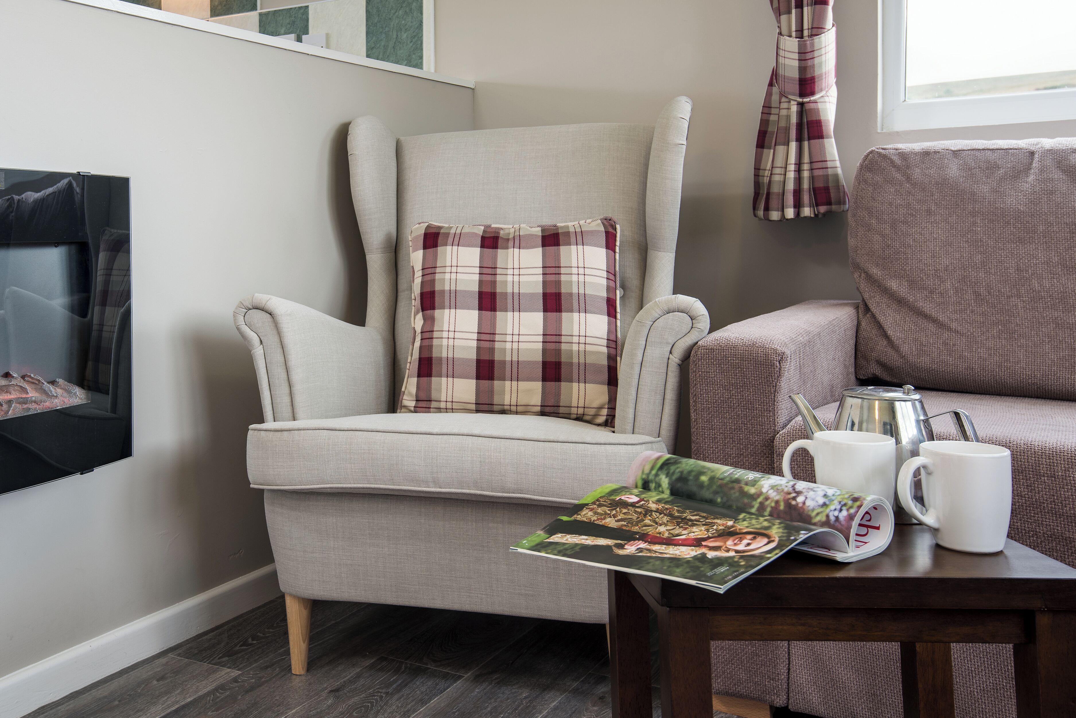 Un coin salon confortable avec un fauteuil à haut dossier, un coussin à carreaux et une table sur laquelle se trouvent un magazine, une tasse et une théière.