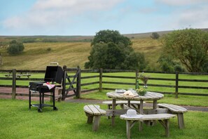A picnic setup with a grill, table, and benches in a grassy area with a view of rolling hills and trees. - Brown Rigg Lodges (Hexham)
