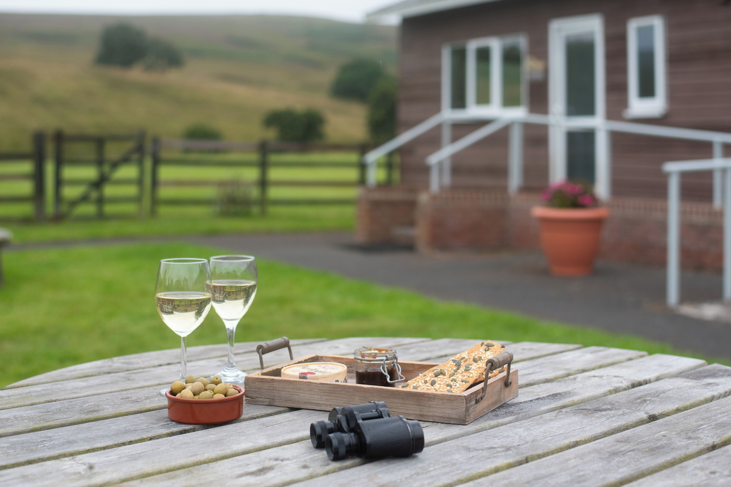 A picnic setup with wine glasses, olives, cheese, and bread on a wooden table with a scenic view of a house and hills in the background.