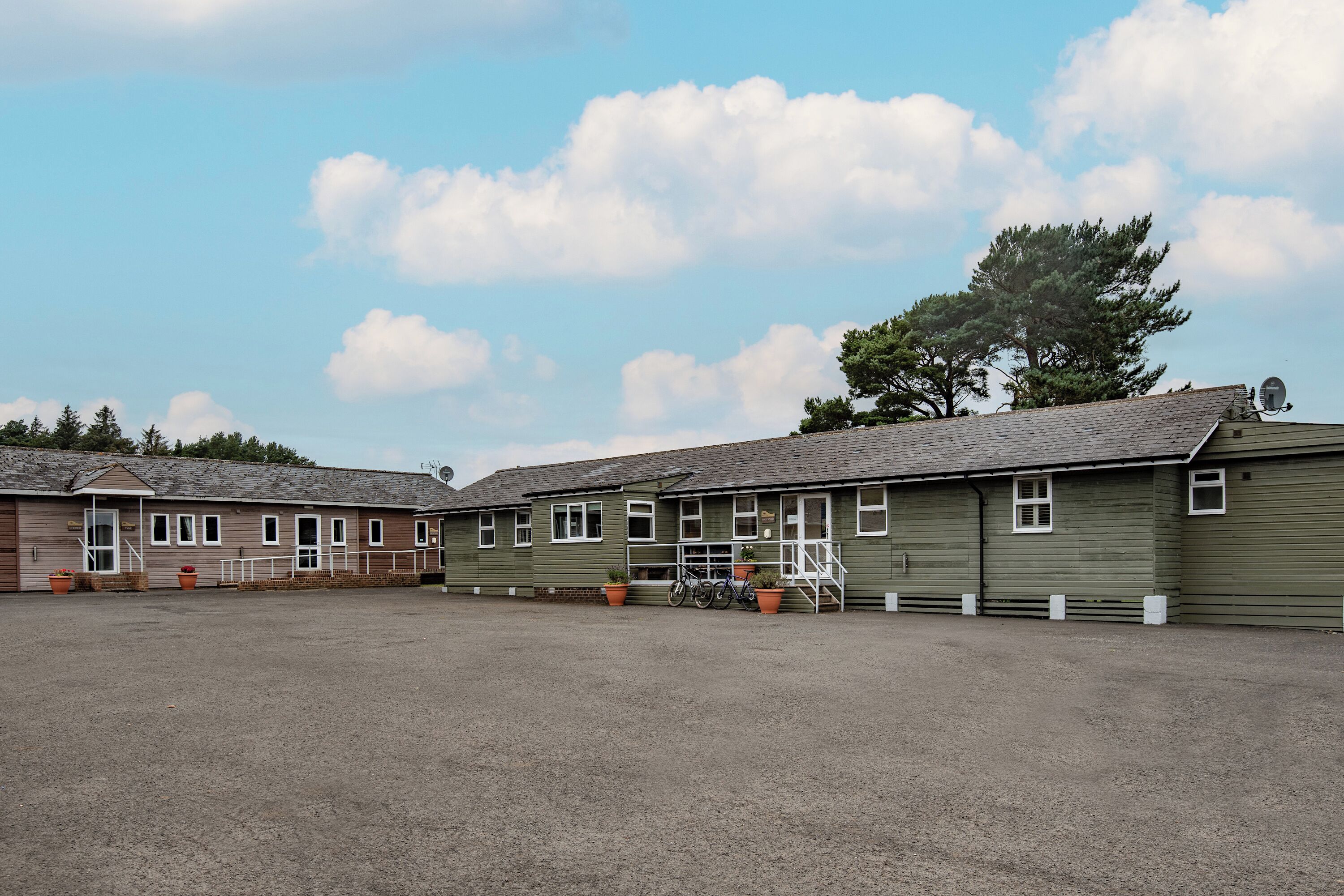 A row of green buildings with a gravel courtyard, windows, and a tree in the background.
