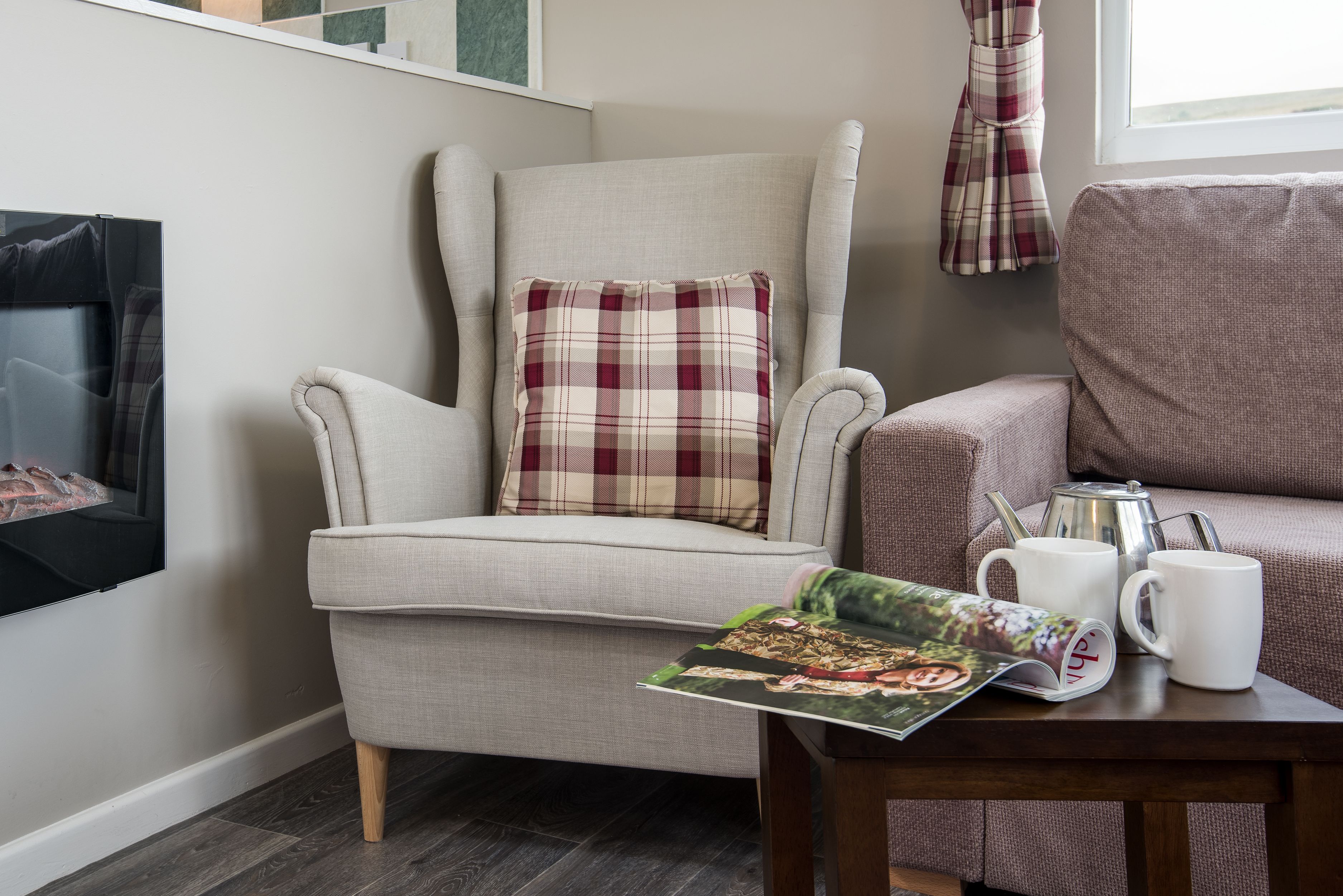 Un salon chaleureux avec un fauteuil à haut dossier, un coussin à carreaux, un magazine, une cafetière et deux tasses blanches posées sur une table en bois.