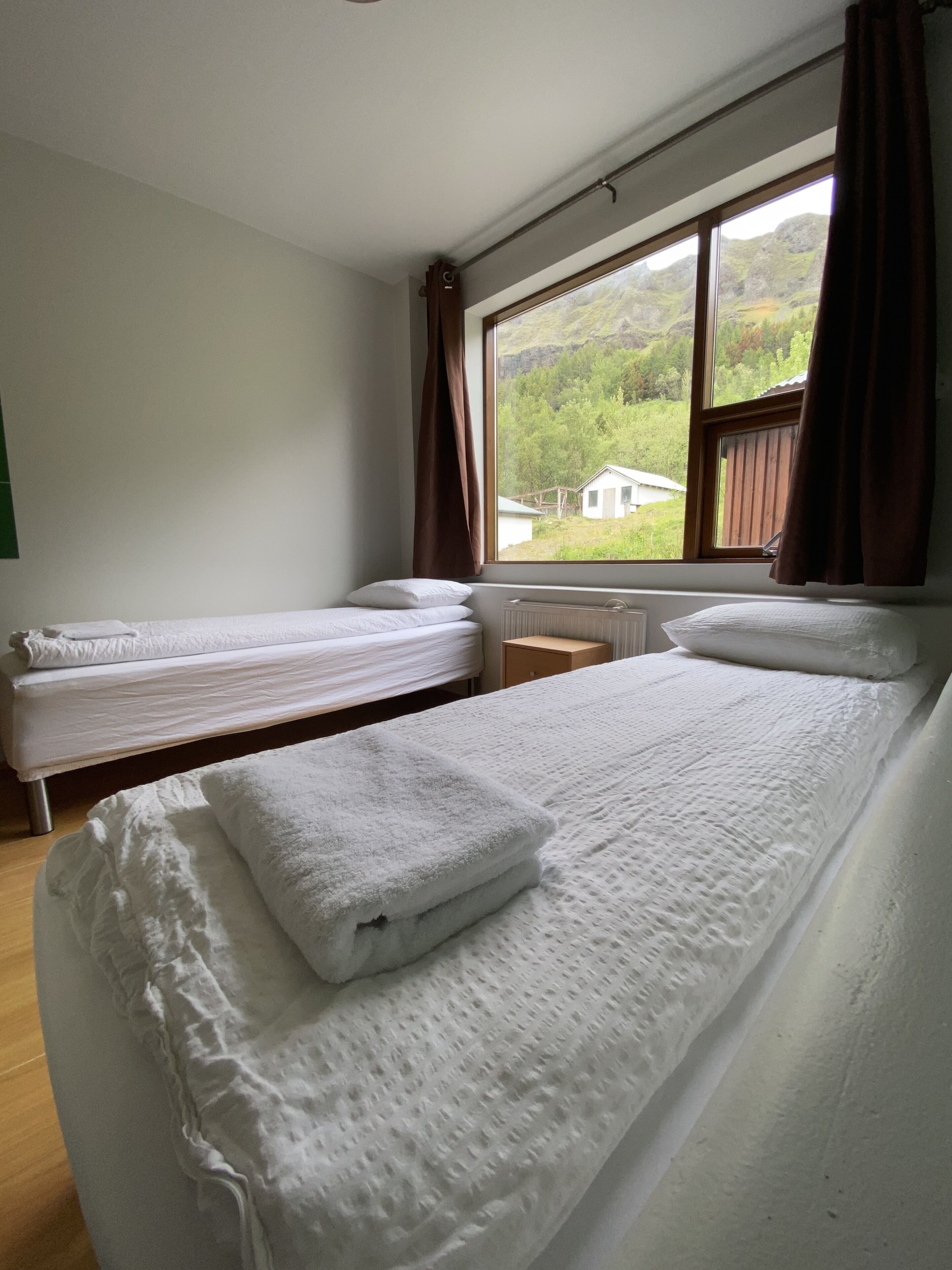 A room with two beds, a window with curtains, and a view of greenery outside.