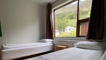 A room with two beds, a window with curtains, and a view of greenery outside.