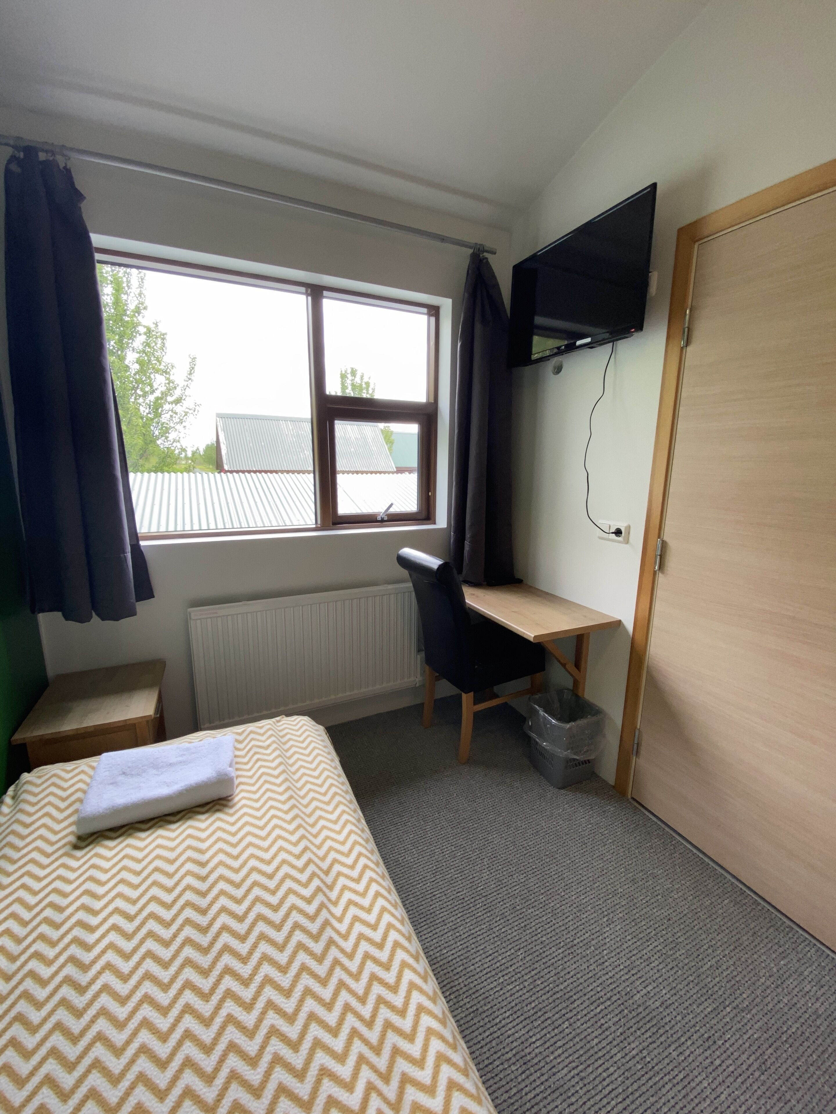 Habitación de hotel con cama, escritorio, silla, televisor montado en la pared y ventana con cortinas.