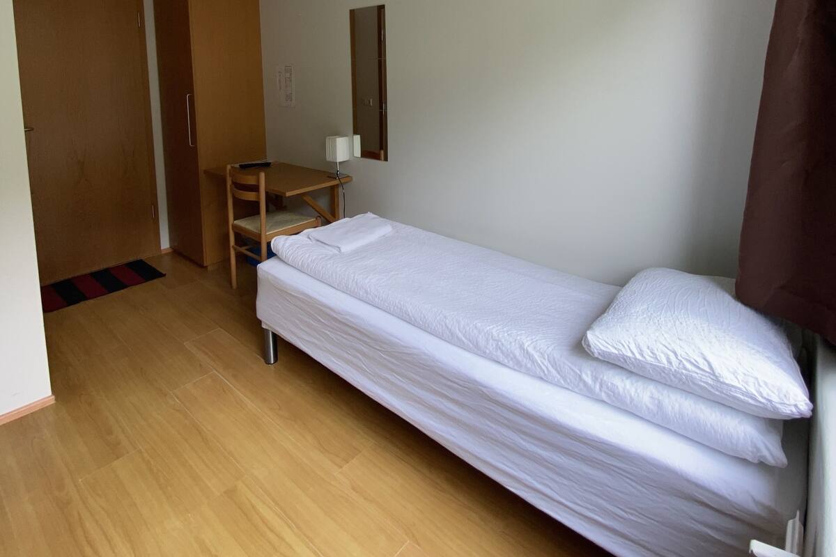 Ein kleines Zimmer mit einem Einzelbett, einem Holztisch, einem Stuhl und einem Spiegel an der Wand.