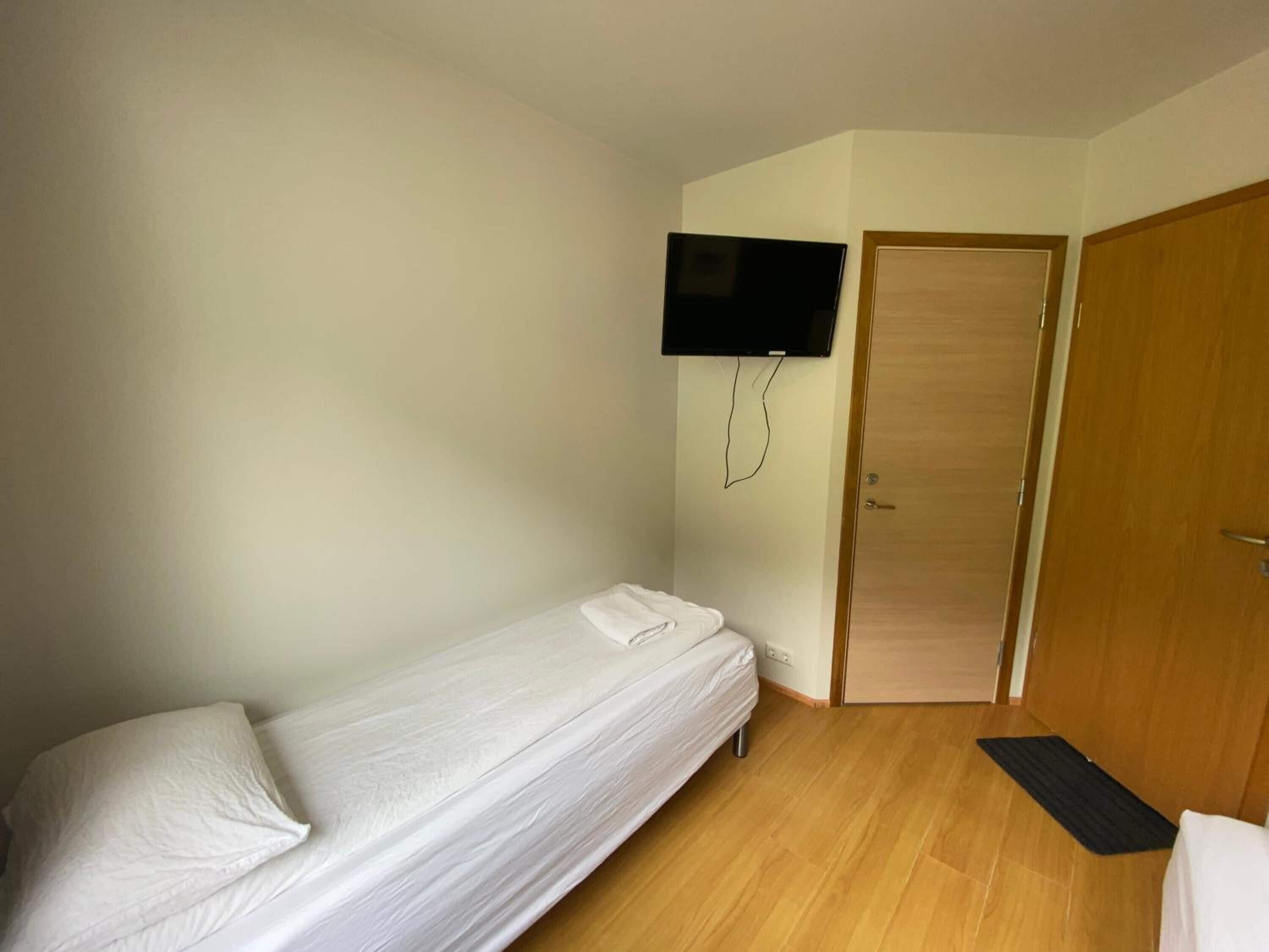 Una habitación pequeña con cama individual, suelo de madera, cama blanca, televisor montado en la pared y puerta de madera.