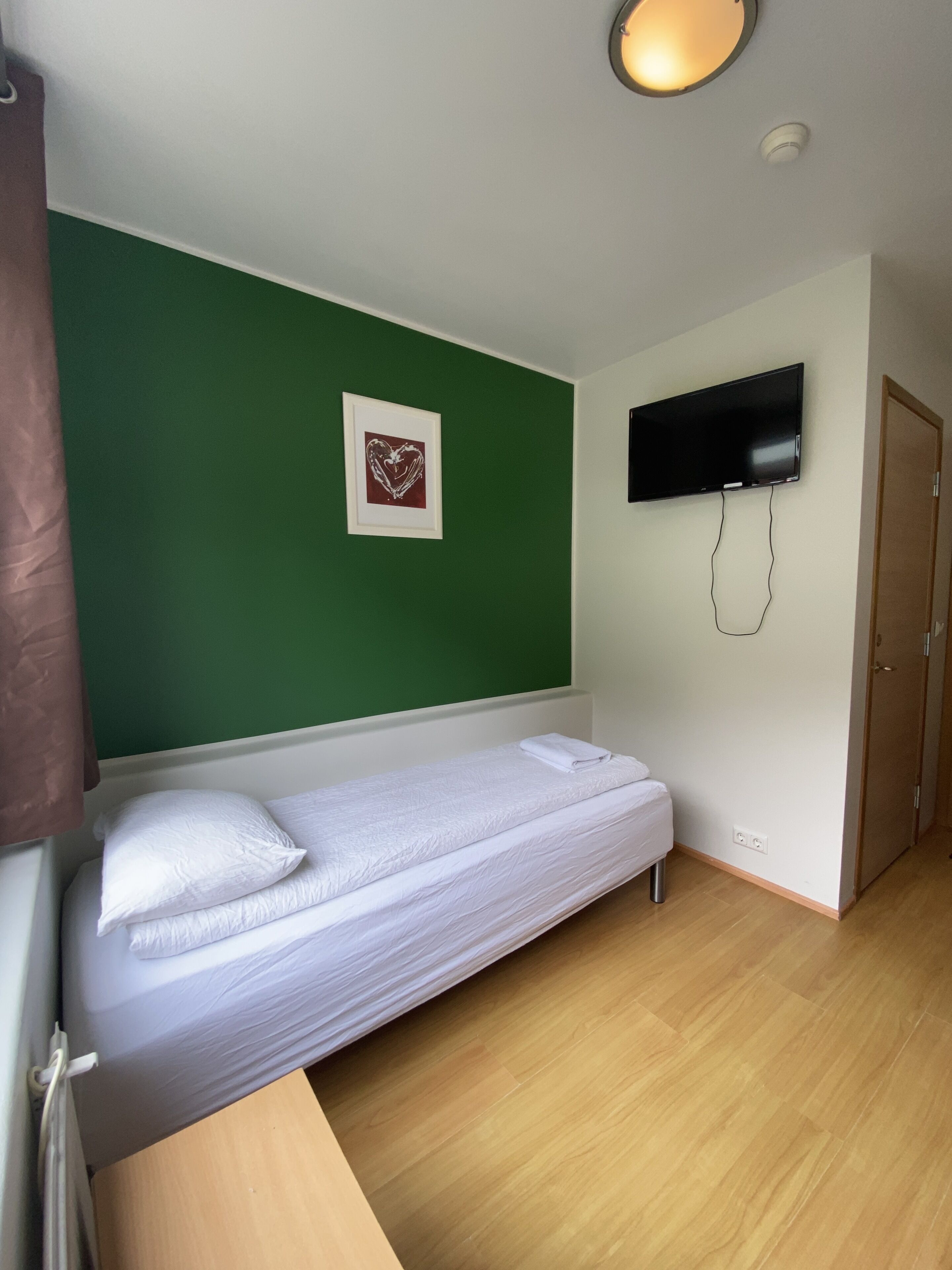 A small, single-bed room with a green wall, a mounted TV, and a wooden floor.