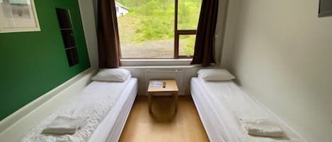 A room with two single beds, a small table, a window with curtains, and a green wall.