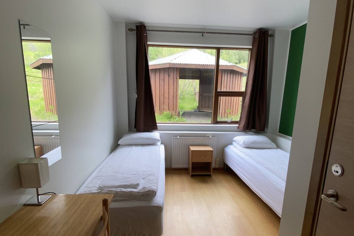 Ein Zimmer mit zwei Einzelbetten, einem Holztisch, einem Stuhl, einem Heizkörper und einem Vorhangfenster.