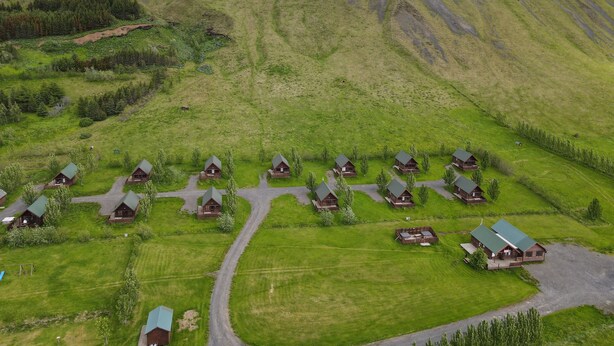 Een groepje blokhutten met groene daken, gerangschikt in een halve cirkel rondom een centrale weg, met op de achtergrond ruige bergen en een begrazend landschap.
