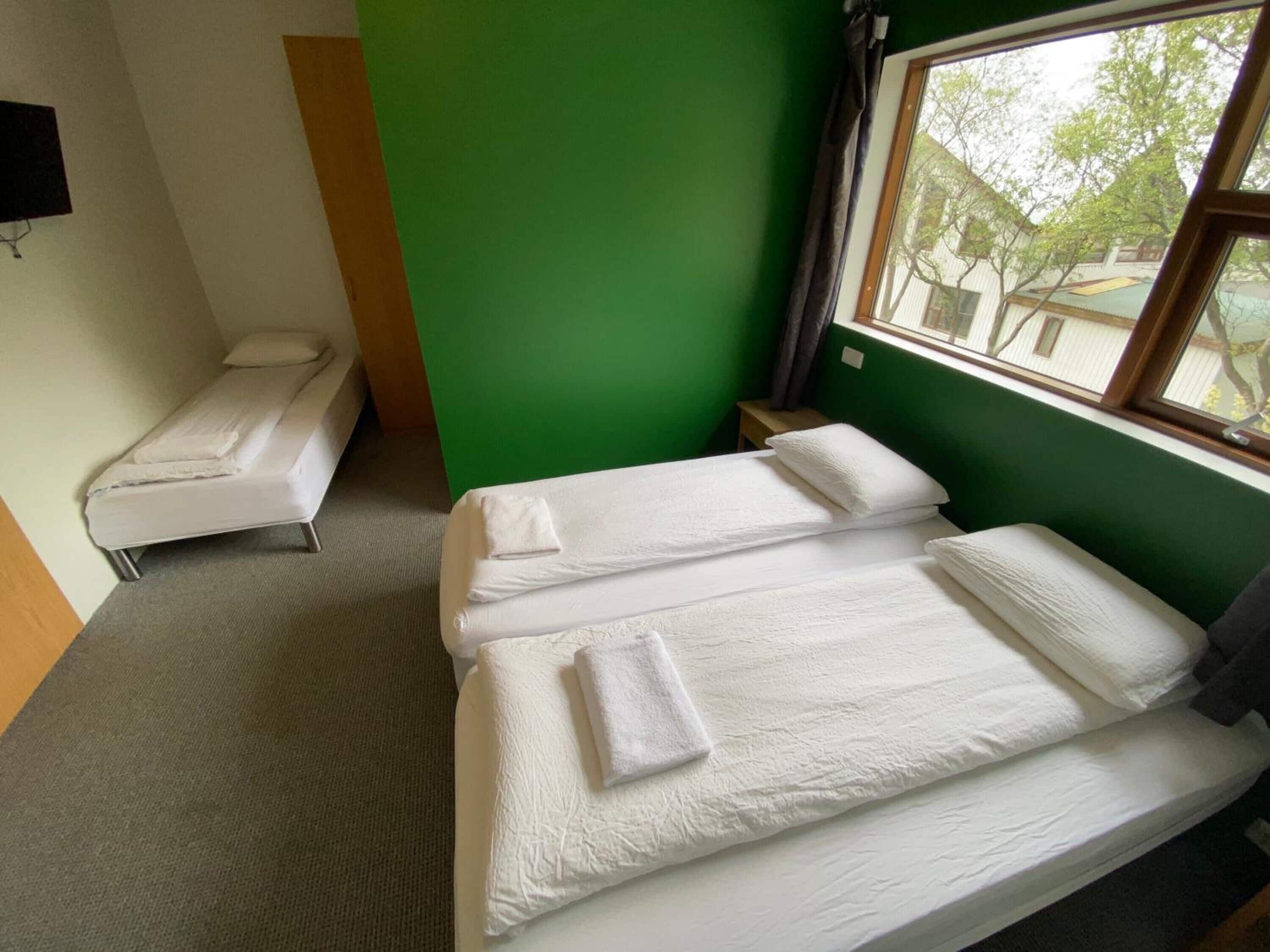 Ein Zimmer mit zwei Einzelbetten, einer grünen Wand, einem Fenster mit Blick auf Gebäude und einem kleinen Tisch mit Lampe.