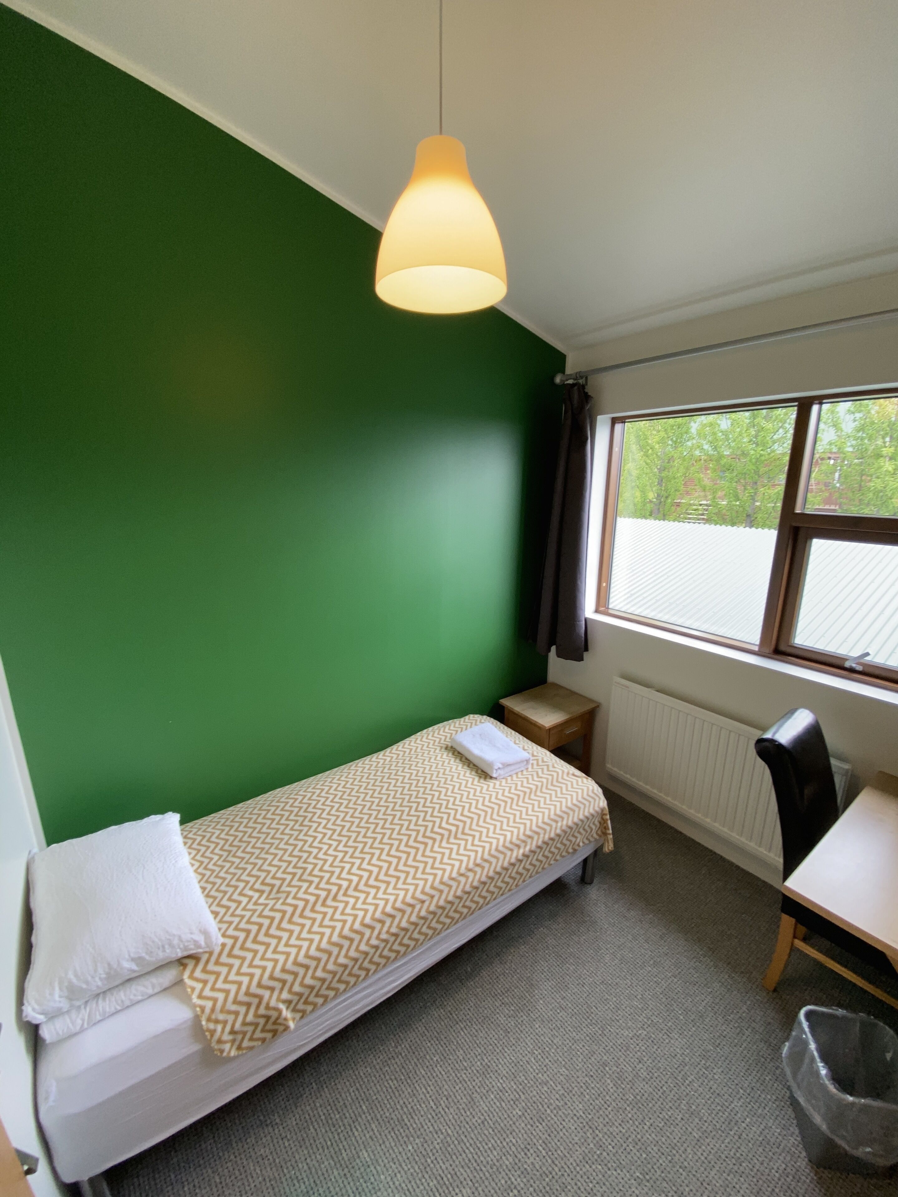 A small, single-bed room with a green wall, a window with blinds, a desk, and a chair.