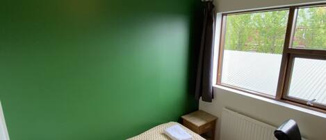 Una habitación pequeña con una cama individual, pared verde, ventana con persianas, escritorio y una silla.
