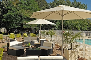 An outdoor seating area with wicker furniture and umbrellas, a pool in the background, and a white fence surrounding the space. - Logis Hotel La Promenade (Coings)