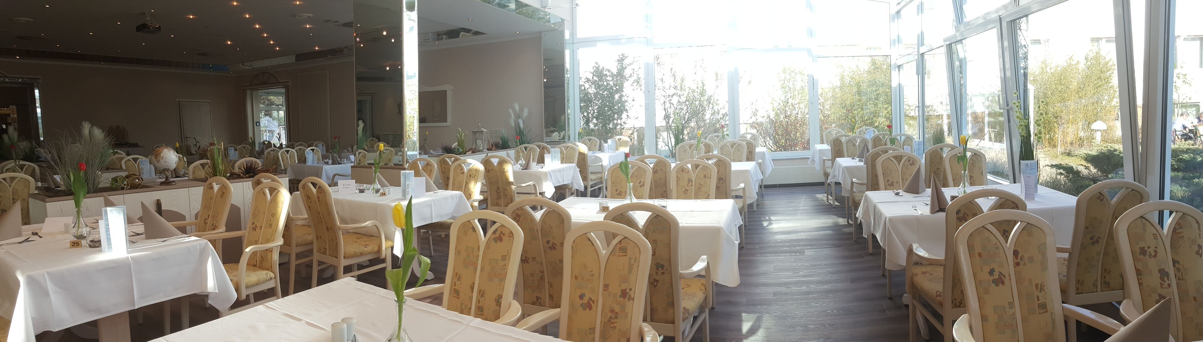 A restaurant with multiple tables and chairs, large windows, and a view of greenery outside.