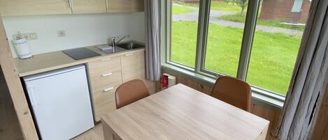 A compact kitchen with wooden cabinets, a sink, and a small refrigerator. There is a dining table with four chairs and a window with a view of the outdoors.