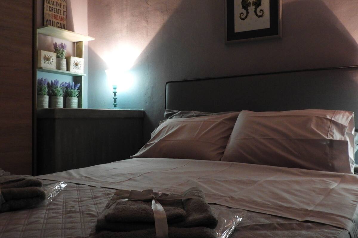 A neatly made bed with pillows and a blanket, a bedside table with a lamp, a shelf with decorative items, and a framed picture on the wall.