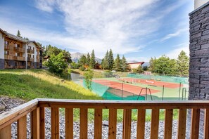 A tennis court with a wooden deck in the foreground, surrounded by a stone wall and a building with balconies. - Sowell Residences Pra Loup (Uvernet-Fours)