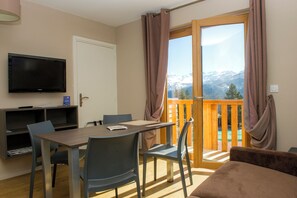 A room with a dining table, chairs, a TV, and a view of mountains through a large window. - Sowell Residences Pra Loup (Uvernet-Fours)