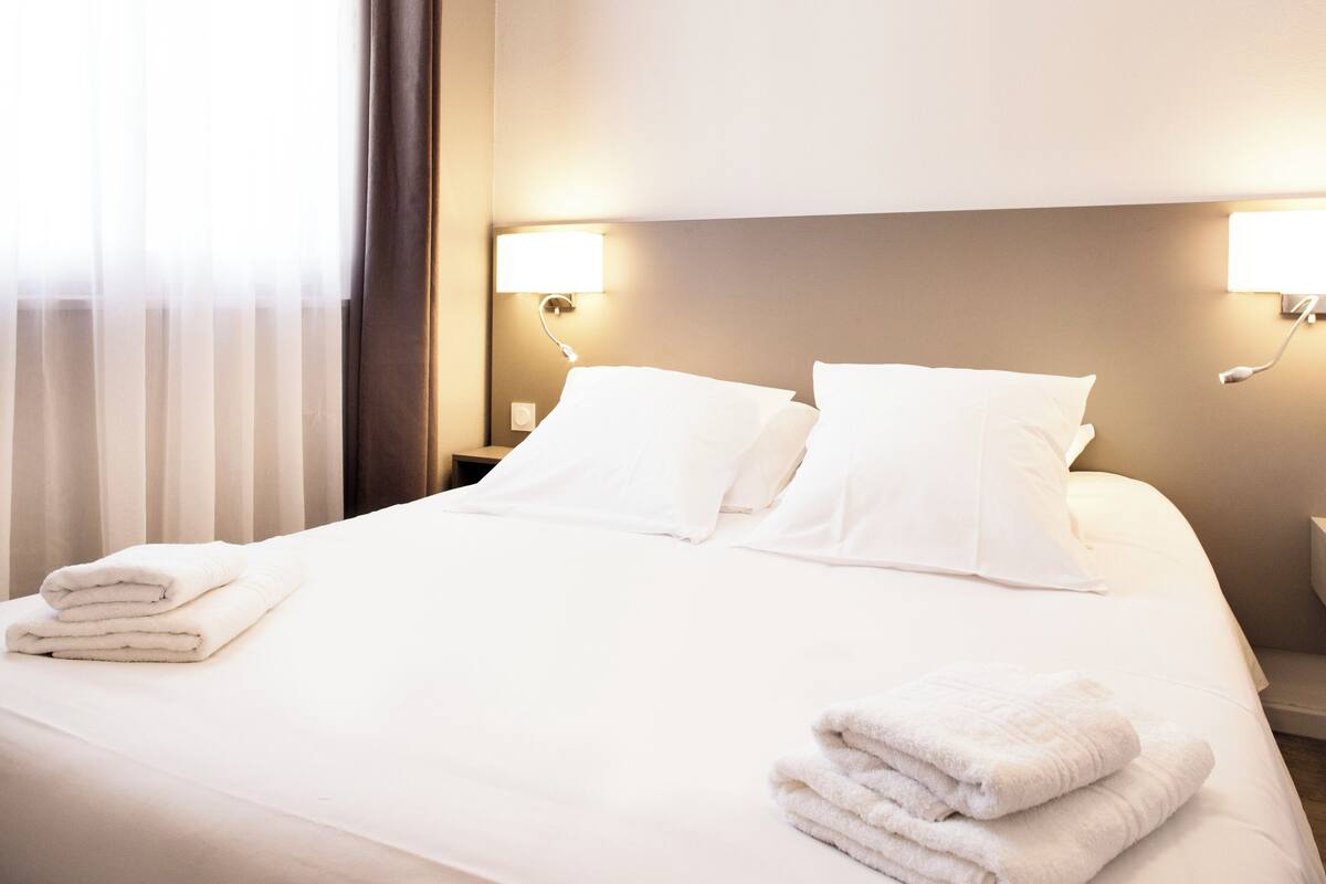 A hotel room with a neatly made bed, two bedside lamps, and a window with curtains.