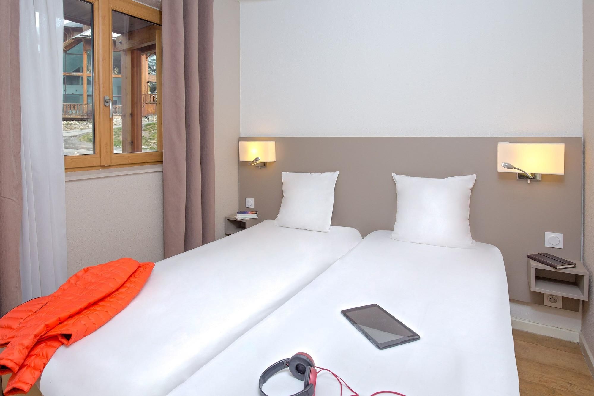 Une chambre d’hôtel avec deux lits, une fenêtre avec des rideaux, des lampes de chevet, une table de chevet avec un livre et une couverture sur l’un des lits.