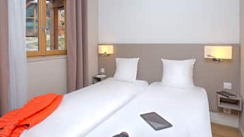 Een hotelkamer met twee bedden, een raam met gordijnen, nachtlampjes, een nachtkastje met een boek, en een dekbed op een van de bedden.