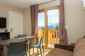 A room with a dining table, chairs, a TV, and a balcony with curtains. - Sowell Residences Pra Loup (Uvernet-Fours)