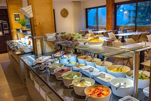 A buffet with various food items and utensils, a clock on the wall, and a view of the outdoors through large windows. - Sowell Residences Pra Loup (Uvernet-Fours)