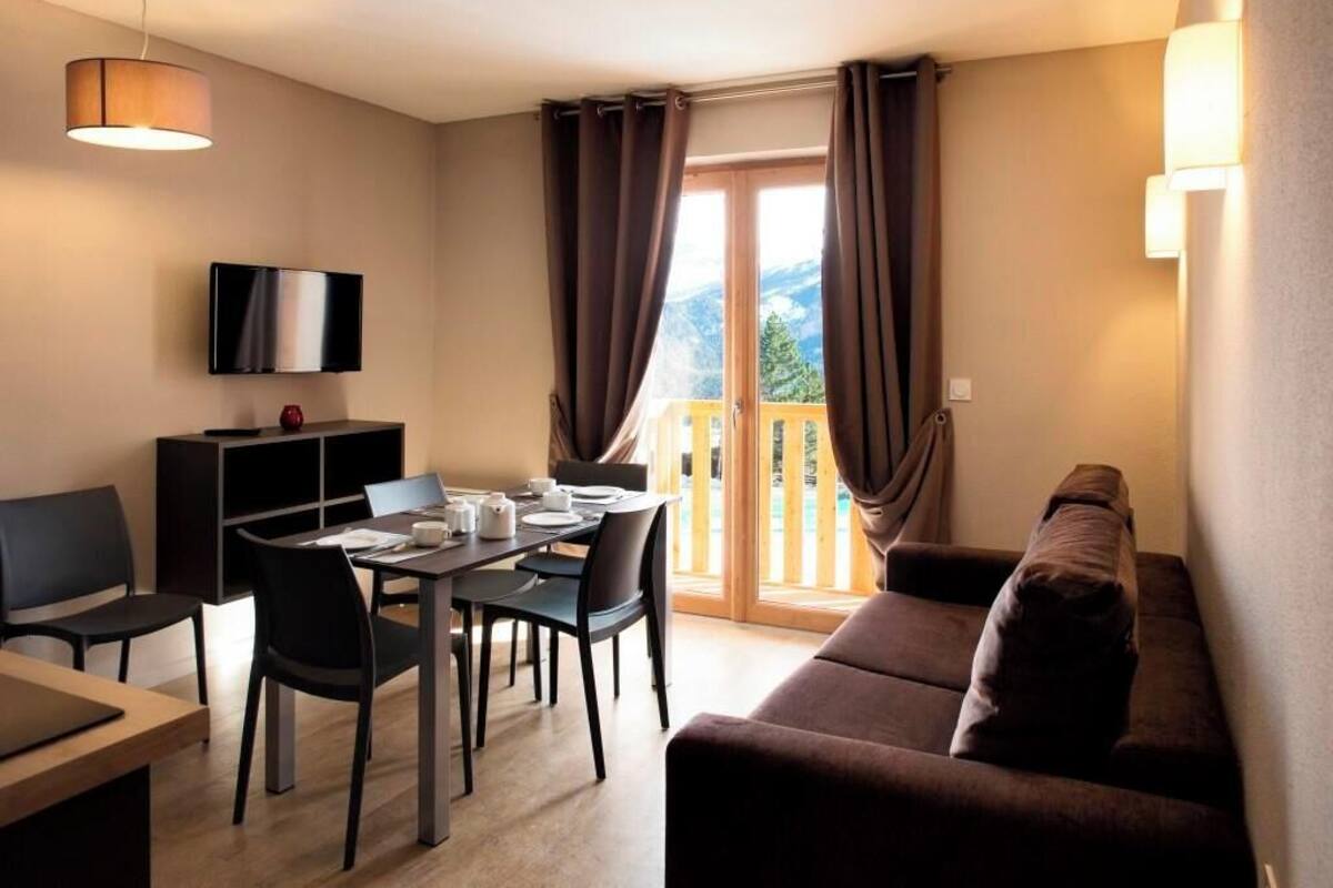 A modern living room with a dining area, a flat-screen TV mounted on the wall, a sofa, and a balcony with curtains.