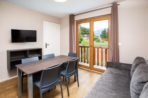 A room with a wooden dining table, chairs, a flat-screen TV, a grey sofa, and a sliding glass door leading to a balcony with a view of greenery and buildings. - Sowell Residences Pra Loup (Uvernet-Fours)
