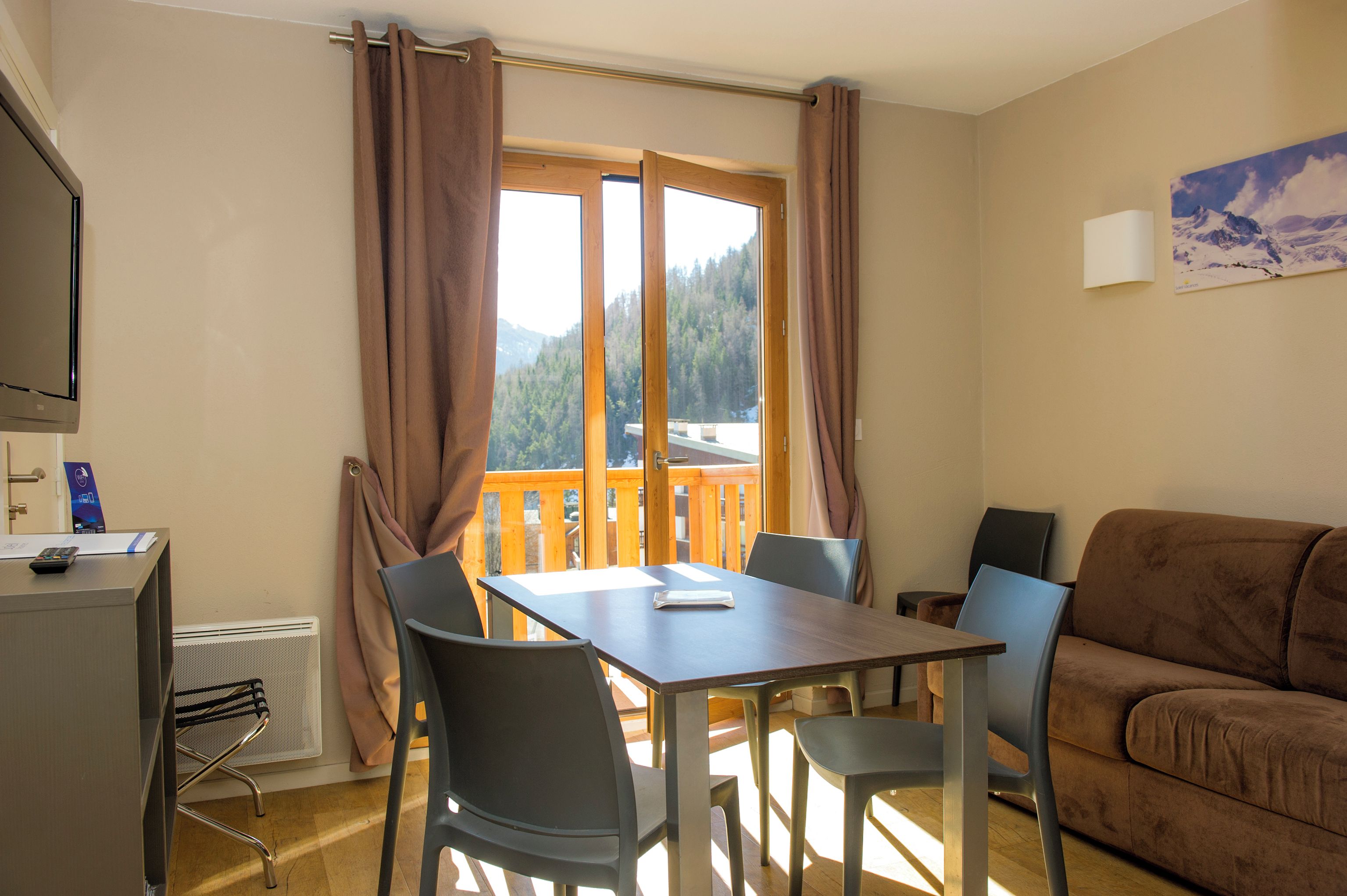 Une pièce comprenant une table et des chaises, un canapé, une fenêtre avec des rideaux, un radiateur et un téléviseur fixé au mur.