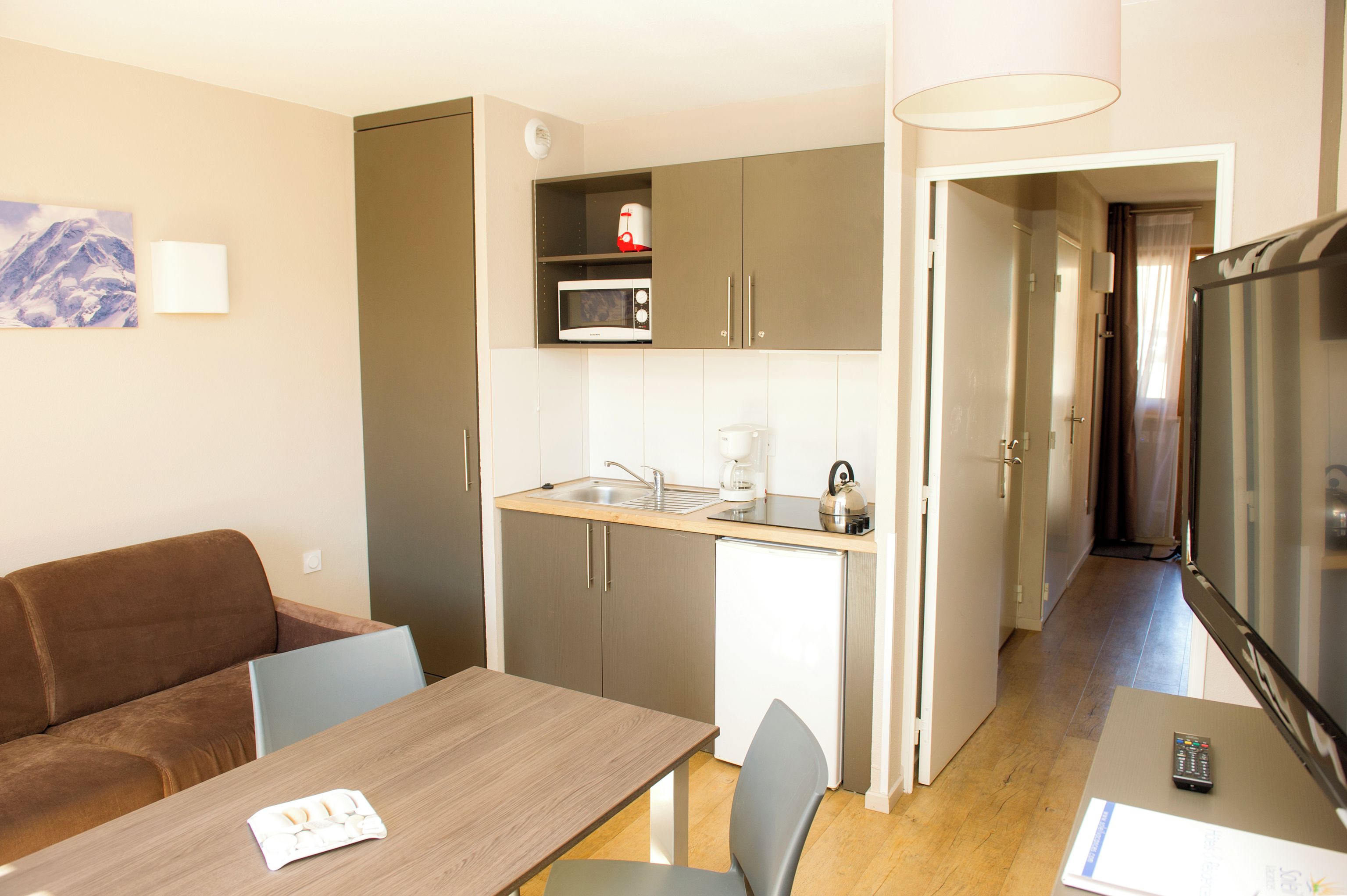 A compact kitchen with a microwave, coffee maker, and kettle, a dining area with a table and chairs, and a living area with a sofa and a TV.