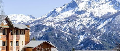 Un chalet en bois, sur lequel on peut lire l’inscription « Hôtel Les Bergers », se dresse devant des montagnes enneigées.