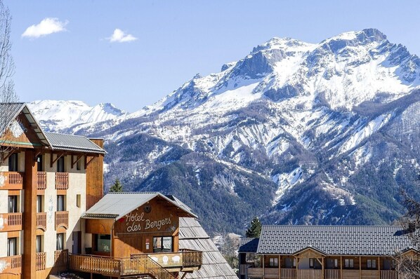 A wooden chalet with a sign reading "Hotel Les Bergers" in front of snow-covered mountains. - Sowell Residences Pra Loup (Uvernet-Fours)