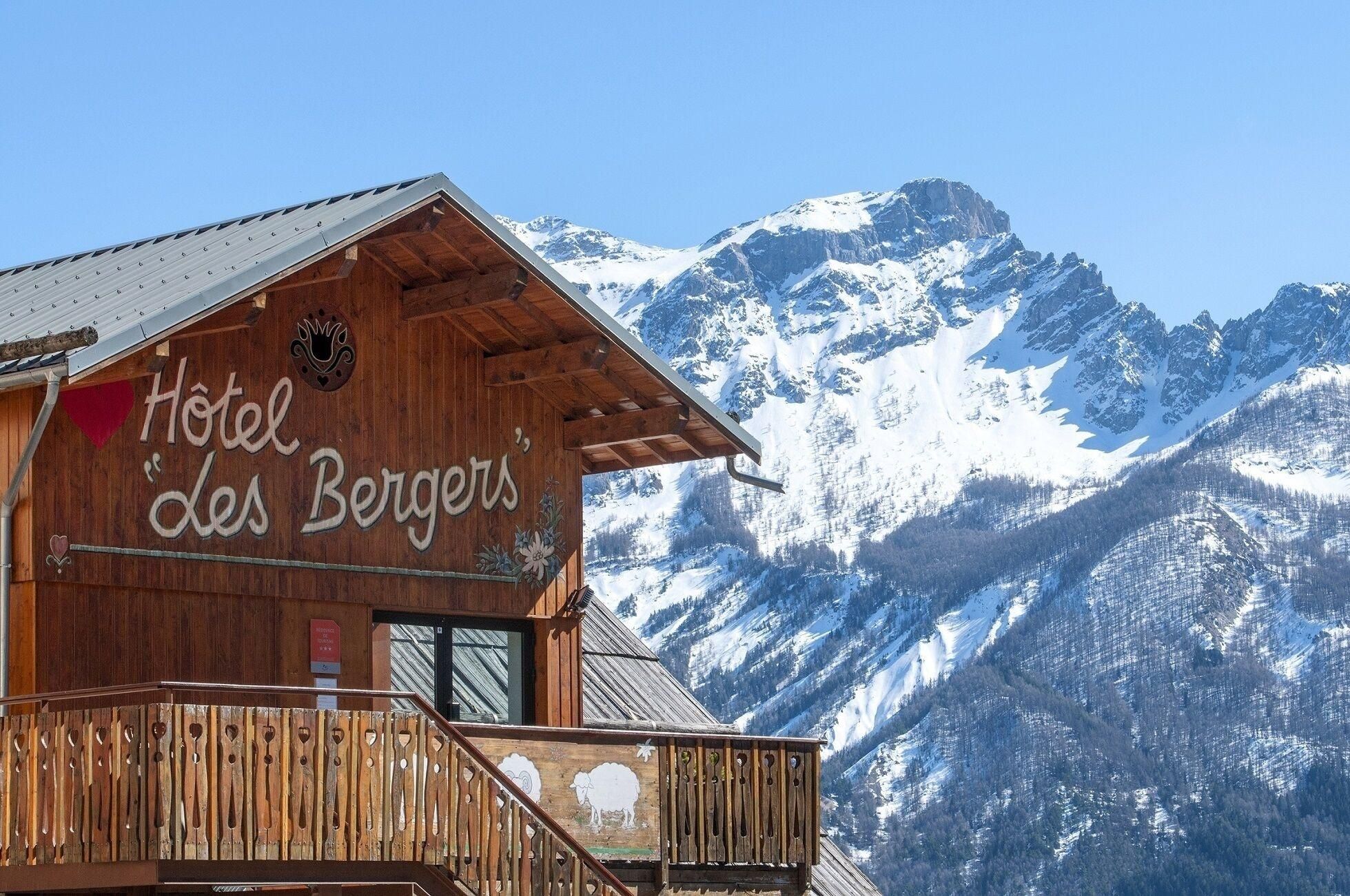 Una cabaña de madera con un cartel que dice "Hotel des Bergers" al fondo de montañas nevadas.