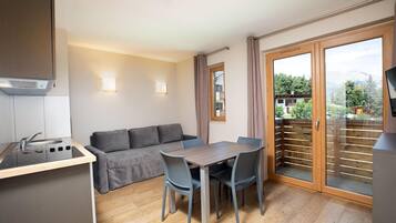Un espacio habitable compacto con kitchenette, un sofá, una mesa de comedor con sillas y un ventanal corredero que da a un balcón.