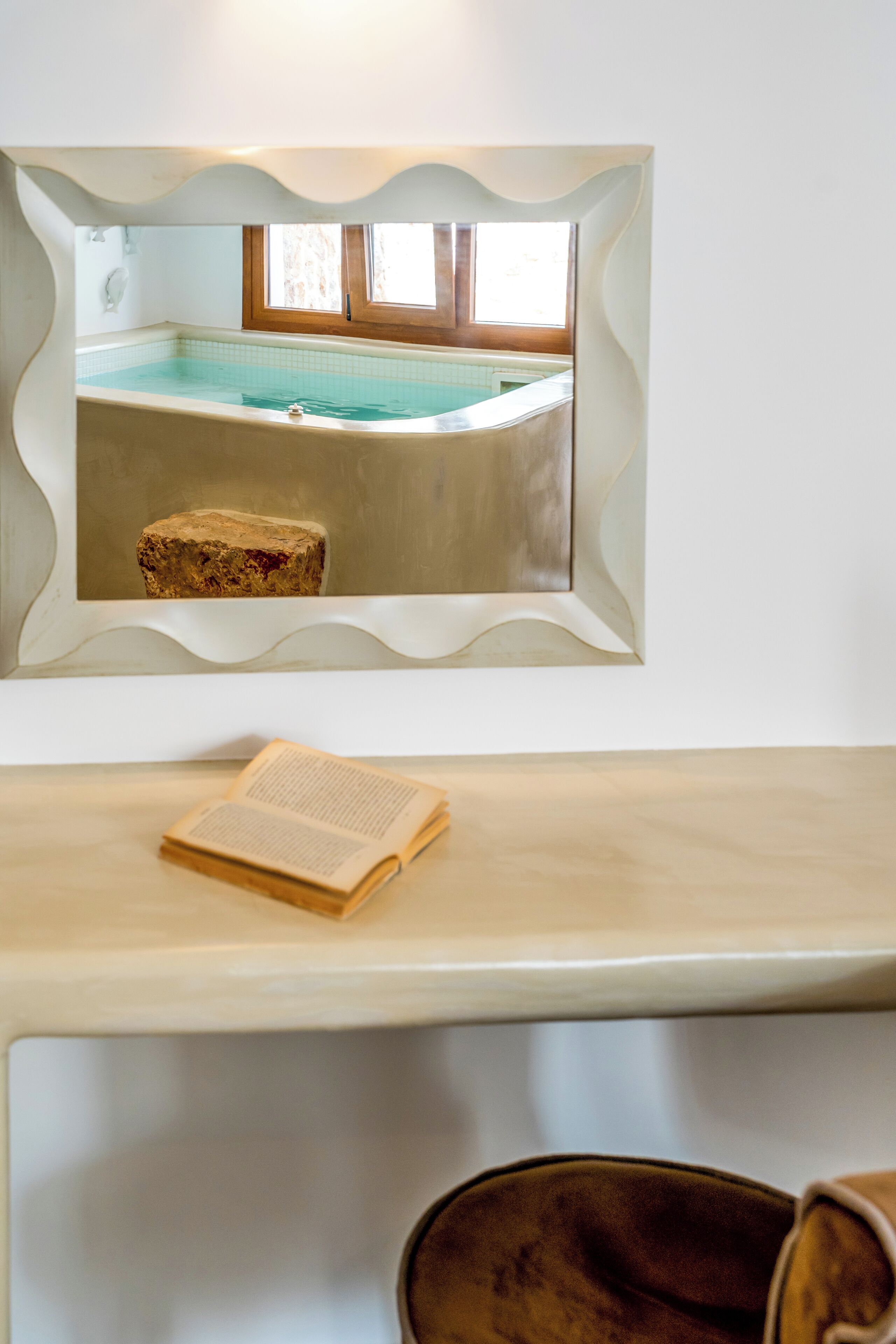 A framed picture of a swimming pool with a wooden stool in front, a book on a table, and a bathtub.