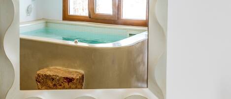 A framed picture of a swimming pool with a wooden stool in front, a book on a table, and a bathtub.