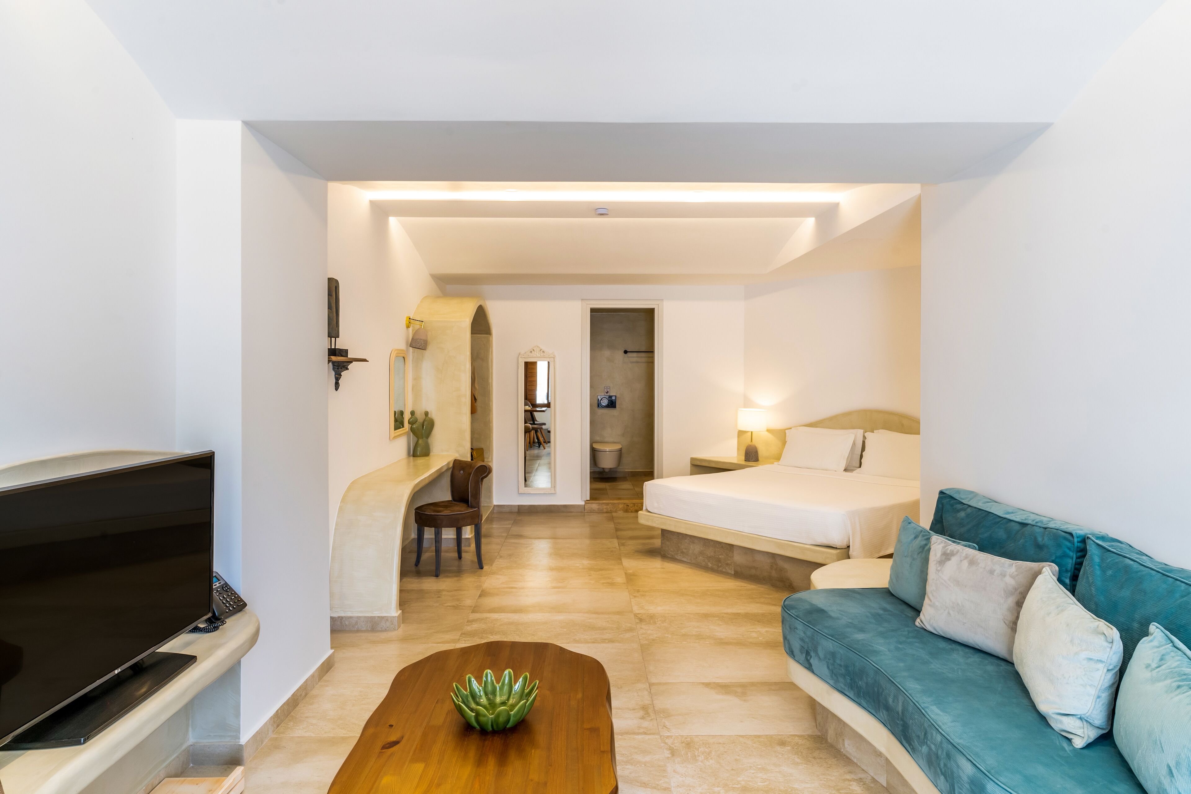A modern hotel room with a flat-screen TV, a wooden coffee table, a teal sofa, and a bed with a nightstand.