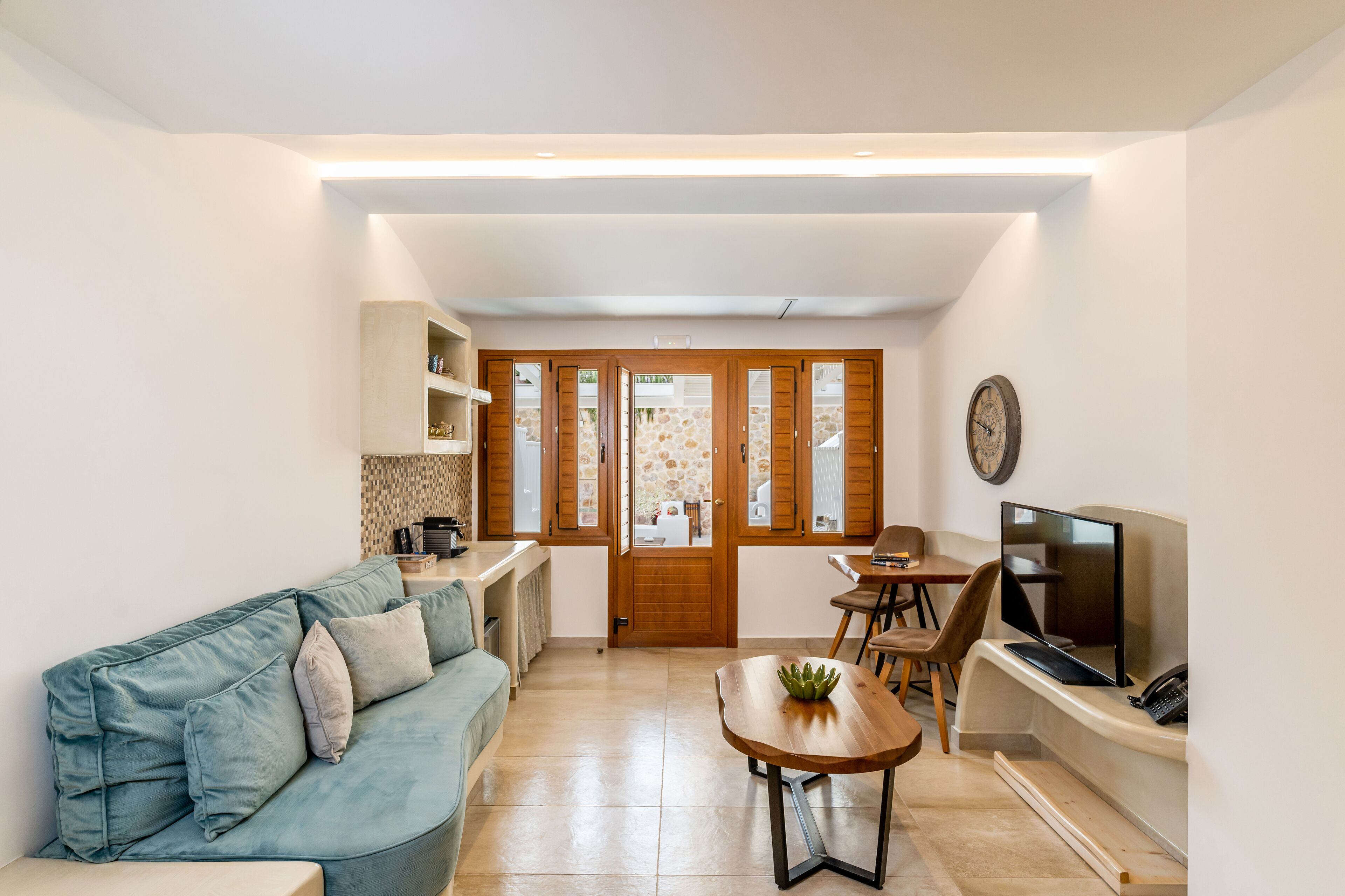 A compact living space with a sofa, a dining table, a TV, and a kitchen area visible through a glass door.