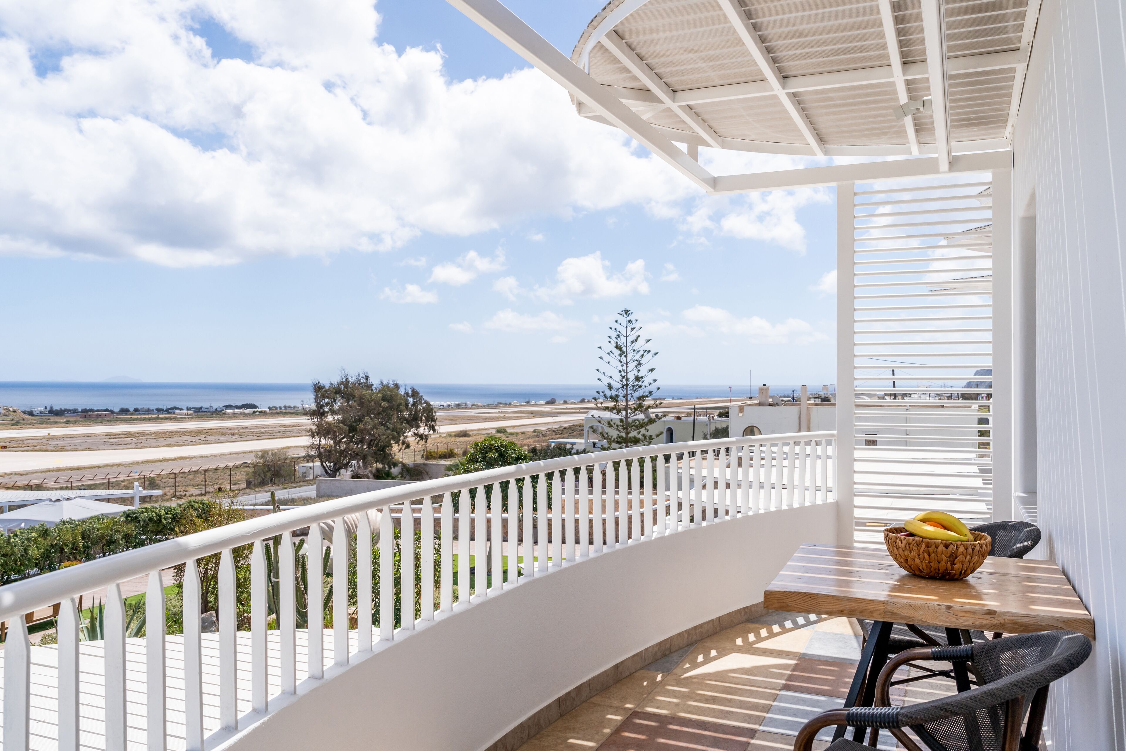 A balcony with a view of the sea, a wicker chair, a table with a fruit bowl, and a white railing.