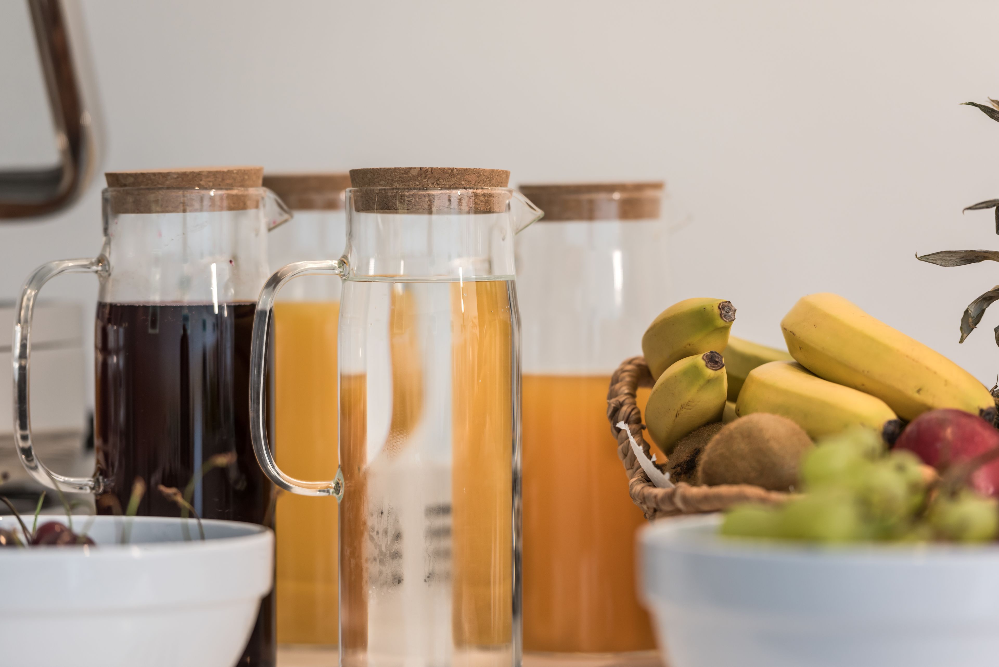 Una encimera de cocina con tres jarras de vidrio que contienen diversas bebidas y un cuenco de fruta fresca.