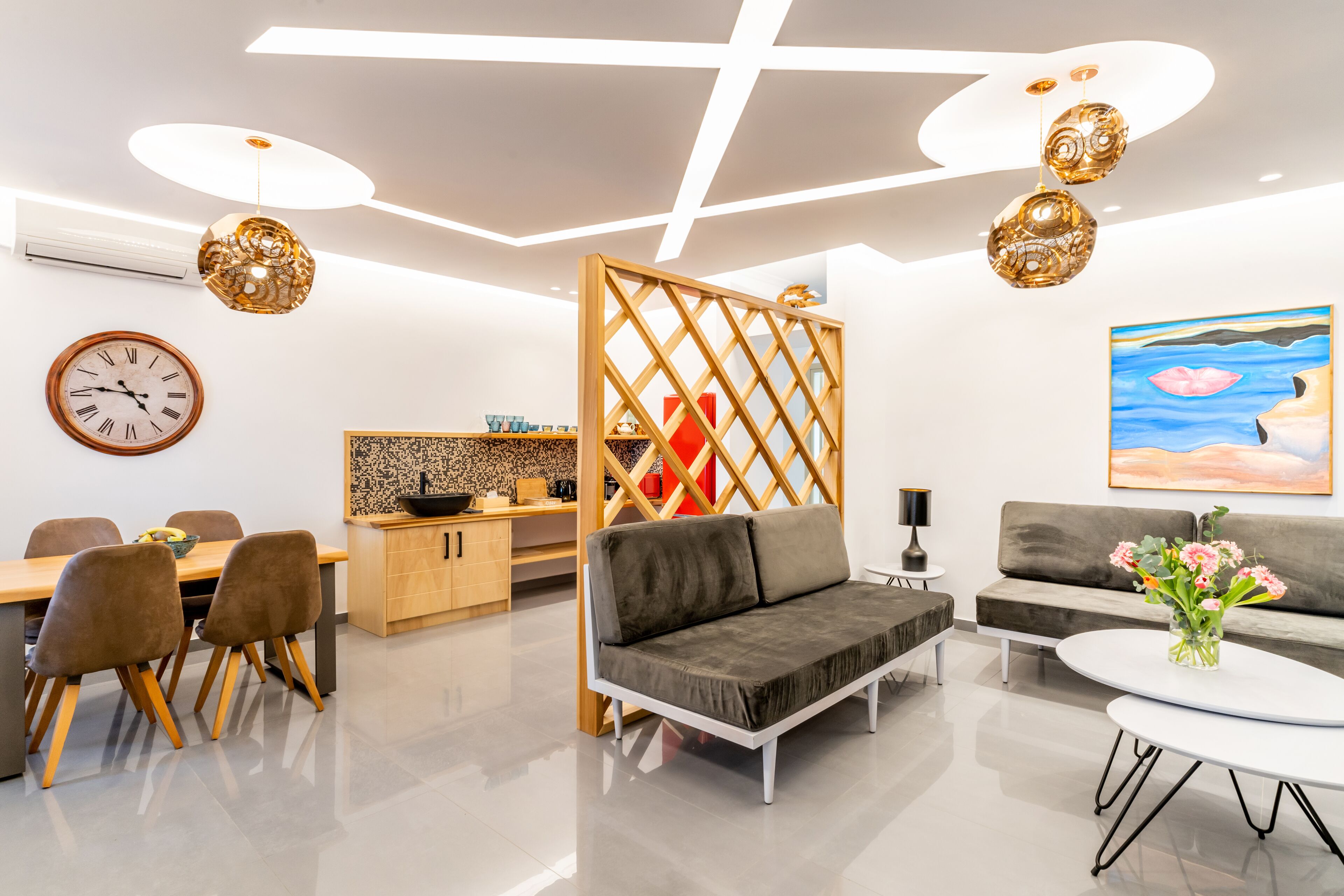A modern living room with a sofa, dining table, and chairs, a kitchen area, and a decorative wall with a clock and artwork.