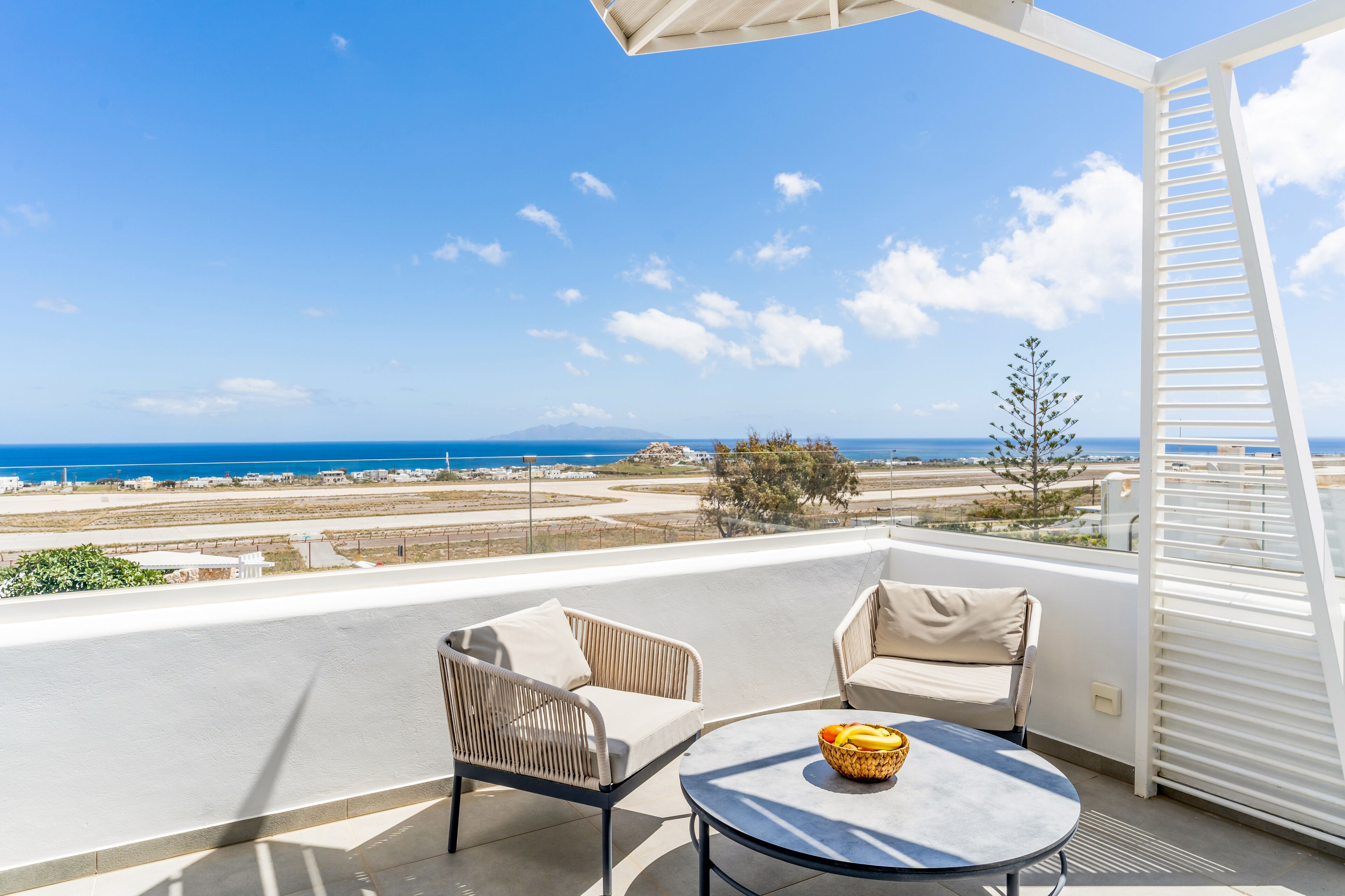 A balcony with two chairs and a table, overlooking a coastal landscape with buildings and the sea.