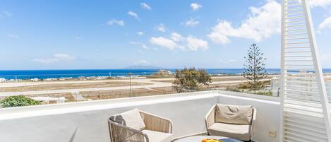 A balcony with two chairs and a table, overlooking a coastal landscape with buildings and the sea.