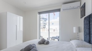 A hotel room with a bed, two rolled towels, a window with a view of buildings, an air conditioner, and a lamp. - San Giljan Stylish Apartment (St. Julian's)
